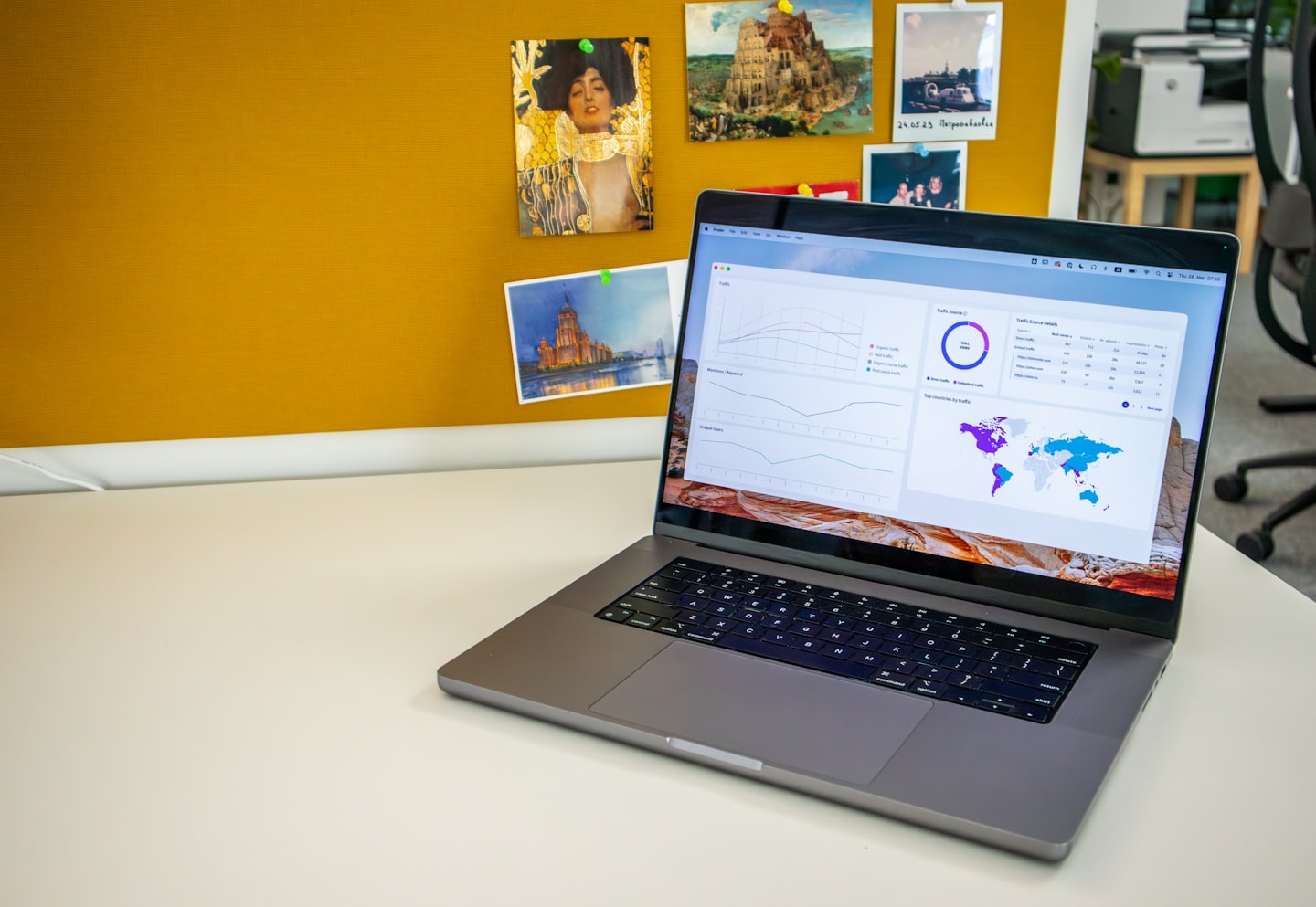 Laptop z wykresami na białym biurku z żółtą tablicą korkową.